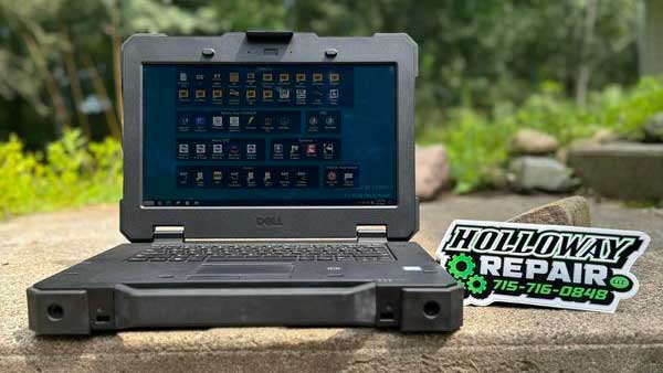 A rugged Dell laptop is open on a stone surface outdoors, displaying a grid of desktop icons. Next to it is a Holloway Repair, LLC business card with a phone number. Mobile Diesel Mechanic