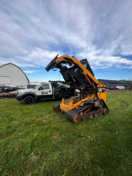 A yellow Caterpillar skid steer loader is parked on grass beside a white utility truck, with an agricultural building in the background under a partly cloudy sky. Mobile Diesel Mechanic